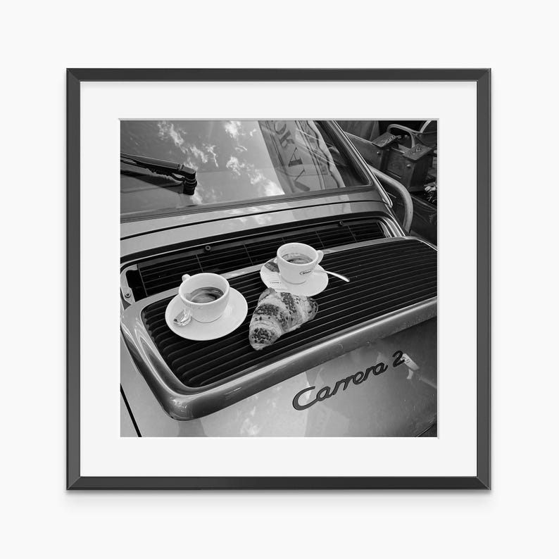 A black and white photograph of a car with two cups of coffee and a croissant on the trunk, framed in a black frame.