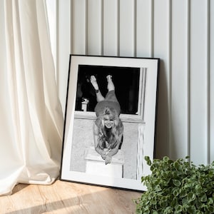 A framed black and white photograph of a woman leaning on a window sill, with a potted plant to the right of the frame.