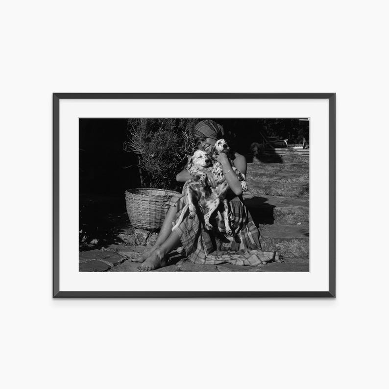 A black and white photograph of a woman sitting on the ground, holding two dogs in her arms.