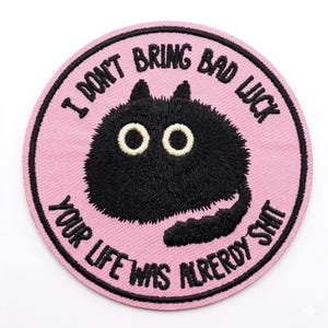 May include: Round embroidered patch with a pink background and a black border. The patch features a black cat with large, round eyes and the text "I DON'T BRING BAD LUCK YOUR LIFE WAS ALREADY SHIT" in a circular pattern.