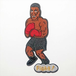 May include: Embroidered patch featuring a cartoon boxer in a fighting stance. The boxer has brown skin, red boxing gloves, and black shorts. The word "Fight!" is embroidered on a cloud-shaped design below the boxer.