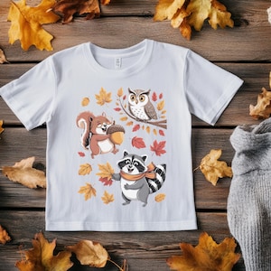 Puede incluir: Camiseta blanca con un diseño de dibujos animados de un búho en una rama, una ardilla sosteniendo una bellota y un mapache con una bufanda sosteniendo hojas. El diseño incluye hojas de otoño en tonos naranja y rojo.