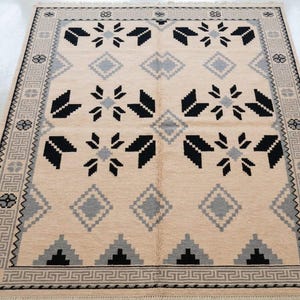 Puede incluir: Alfombra rectangular beige con un diseño geométrico. Presenta formas florales y de diamante en negro y gris, con un borde de patrones grises y negros repetidos. La alfombra es de color neutro y encajaría en muchas habitaciones.