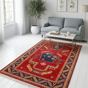 Puede incluir: Una alfombra rectangular roja con un detallado patrón geométrico en azul marino, dorado y verde. La alfombra tiene un borde con flecos y está colocada en un salón con un sofá gris, una mesa de centro y un olivo en maceta.