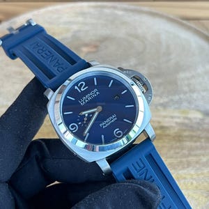 May include: A stainless steel Panerai Luminor Marina Automatic watch with a blue dial and blue rubber strap. The watch face displays the time with luminous hands and markers. The strap is marked with the Panerai logo.