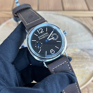 Pode incluir: Um relógio Radiomir Panerai em aço inoxidável com mostrador preto, algarismos e ponteiros brancos e um pequeno subdial de segundos. Possui uma pulseira de couro castanho com costuras visíveis. O relógio é exibido sobre uma superfície de madeira.