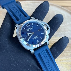 May include: A stainless steel Panerai Luminor Marina automatic watch with a blue dial and blue rubber strap. The watch features a date display and luminous hands and markers. The strap is marked "PANERAI" and "OFFICINE".