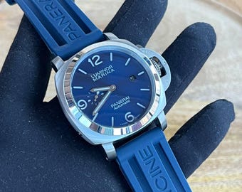 May include: A stainless steel Panerai Luminor Marina automatic watch with a blue dial and blue rubber strap. The watch features a date display and luminous hands and markers. The strap is marked "PANERAI" and "OFFICINE".