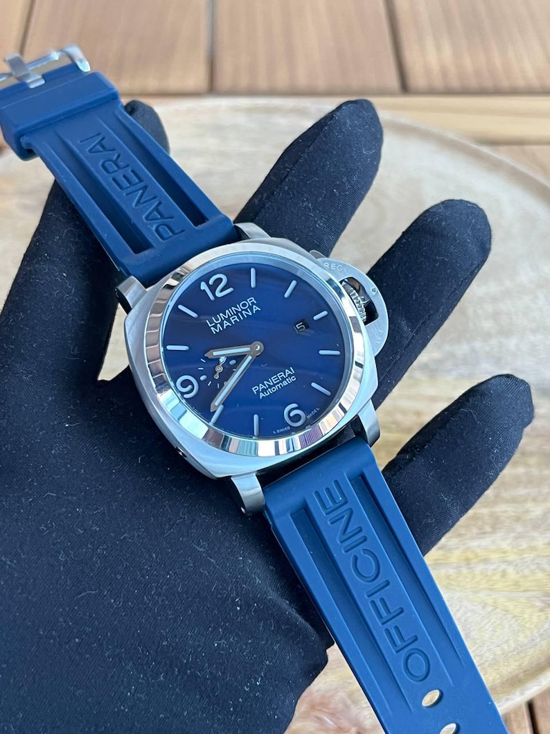 May include: A stainless steel Panerai Luminor Marina automatic watch with a blue dial and blue rubber strap. The watch features a date display and luminous hands and markers. The strap is marked "PANERAI" and "OFFICINE".