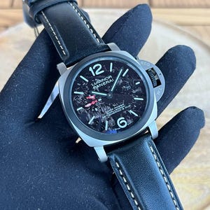 Pode incluir: Um relógio de luxo com pulseira de couro preta e caixa de aço inoxidável. O mostrador do relógio apresenta um design escuro e texturizado com algarismos e ponteiros brancos. O relógio é marcado "Luminor Panerai" e "Swiss Made".