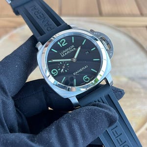 May include: A black Luminor Marina watch with a black rubber strap. The watch face is black with luminous green numerals and hands. The watch has the brand name "PANERAI" on the strap and face.