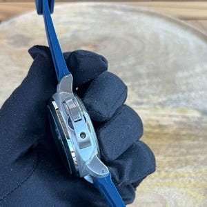 May include: A silver-toned wristwatch with a dark face and a blue band. The watch has a rectangular case and a clasp closure. The band is made of a flexible material. The watch is held by a gloved hand.
