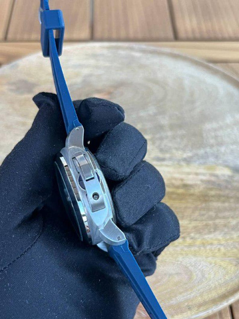 May include: A silver-toned wristwatch with a dark face and a blue band. The watch has a rectangular case and a clasp closure. The band is made of a flexible material. The watch is held by a gloved hand.