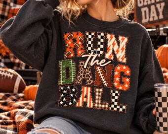 Könnte beinhalten: Ein dunkelgraues Sweatshirt mit Rundhalsausschnitt und dem Schriftzug RUN THE DANG BALL in großen, gemusterten Blockbuchstaben. Die Buchstaben zeigen Football-, Schachbrett-, Blitz-, Leoparden- und Sternmuster in Orange, Weiß und Grün.
