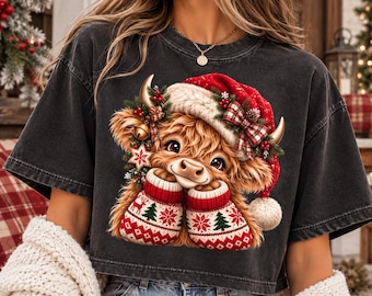 Könnte beinhalten: Ein anthrazitfarbenes Kurzarm-T-Shirt mit einer Grafik eines flauschigen Hochlandrindes, das eine rot-weiße Weihnachtsmütze trägt. Das Rind ist mit Tannenzweigen, einer karierten Schleife und einer Glocke verziert und trägt rot-weiß gemusterte Fäustlinge.