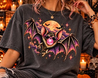 Könnte beinhalten: Ein anthrazitfarbenes T-Shirt mit einer Grafik einer Fledermaus, die eine rosa Sonnenbrille trägt und eine rosa Kaugummiblase bläst. Das Motiv zeigt zudem einen großen gelben Mond, violette Wolken und orangefarbene Sterne auf dem Shirt.
