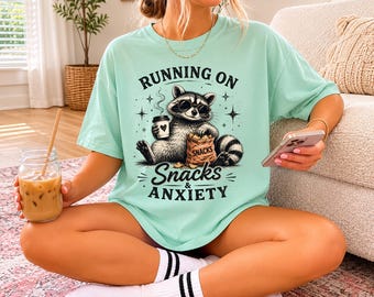 Könnte beinhalten: Hellgrünes T-Shirt mit dem Text "RUNNING ON SNACKS & ANXIETY" und einer Grafik eines Waschbären mit Brille, der eine Kaffeetasse und eine Snacktüte hält. Das Shirt hat eine lockere Passform.
