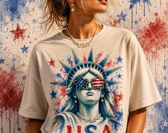 Könnte beinhalten: Ein cremefarbenes Kurzarm-T-Shirt mit einem Grafikdruck der Freiheitsstatue, die sternförmige Sonnenbrillen mit US-Flaggenmustern trägt. Das Design enthält blaue und rote Aquarellakzente sowie den Schriftzug USA in roten Buchstaben.