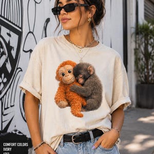 Viral lonely monkey Comfort Colors shirt featuring a baby monkey hugging a stuffed toy, modeled by a stylish couple wearing matching oversized graphic tees in pepper and ivory colors.