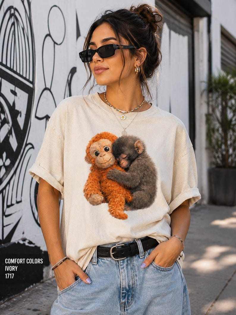 Viral lonely monkey Comfort Colors shirt featuring a baby monkey hugging a stuffed toy, modeled by a stylish couple wearing matching oversized graphic tees in pepper and ivory colors.