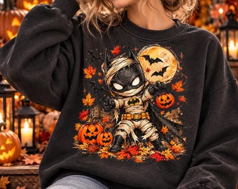 Könnte beinhalten: Ein anthrazitfarbenes Sweatshirt mit Rundhalsausschnitt und einem Grafikdruck eines als Mumie verkleideten Charakters mit Fledermausmaske, der einen Kürbiseimer hält. Das Motiv zeigt orangefarbene Herbstblätter, Fledermäuse und zwei Kürbisse.