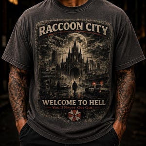 May include: Dark gray t-shirt with a distressed graphic featuring the text "Raccoon City" above a gothic cityscape and the words "Welcome to Hell". The design includes a full moon and an umbrella logo.