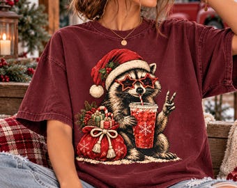 Könnte beinhalten: Ein dunkelrotes Kurzarm-T-Shirt mit einer Grafik eines Waschbären, der eine Weihnachtsmütze und sternförmige Sonnenbrillen trägt. Der Waschbär hält ein rotes Getränk und zeigt ein Friedenszeichen neben einem Geschenkesack.