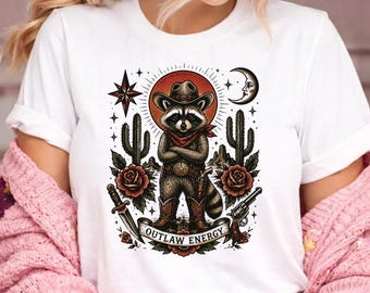 Könnte beinhalten: Weißes T-Shirt mit einem Waschbär-Cowboy-Design. Der Waschbär trägt einen Cowboyhut, ein rotes Halstuch und Stiefel, mit einer Pistole und einem Messer. Das Design umfasst Kakteen, Rosen, einen Stern und den Text "OUTLAW ENERGY".
