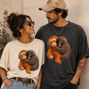 Young couple wearing matching Comfort Colors graphic t-shirts featuring a viral lonely baby monkey hugging a plush toy. Vintage oversized aesthetic shirts in ivory and pepper colors photographed in a warm lifestyle setting, perfect for Etsy apparel listings and animal lover gifts.