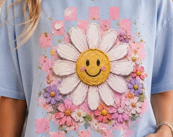 Könnte beinhalten: Ein hellblaues T-Shirt mit einer zentralen Grafik einer großen weißen Margerite mit einem gelben Smiley-Gesicht in der Mitte. Das Design umfasst einen rosafarbenen karierten Hintergrund und kleinere rosa, violette und weiße Blumen, die alle mit einem strukturierten, pastosen Farbeffekt dargestellt sind.