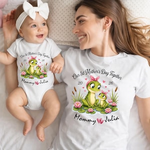 Può includere: Magliette bianche e un body per neonati con una grafica a forma di rana. Il design include il testo "Our 1st Mother's Day Together 20 26 Mommy Julia" con illustrazioni di rane, ninfee e accenti rosa. Un bambino e una donna indossano gli abiti abbinati.