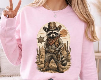 Könnte beinhalten: Hellrosa Sweatshirt mit einem Cartoon-Waschbären im Cowboy-Outfit. Der Waschbär trägt einen braunen Hut, eine Weste und Stiefel, ein rotes Halstuch und eine Pistole. Das Design umfasst einen Sheriff-Stern, Kakteen und einen Wüsten-Sonnenuntergang.