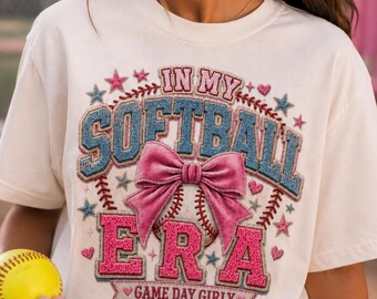 Könnte beinhalten: Ein cremefarbenes Kurzarm-T-Shirt mit einem Grafikdruck, der den Text "IN MY SOFTBALL ERA" und "GAME DAY GIRLY" zeigt. Das Design umfasst einen Softball, eine große rosa Schleife, blau-rosa strukturierte Schriftzüge sowie kleine Sterne.