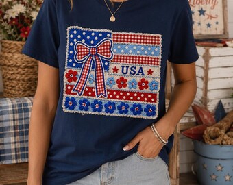 Könnte beinhalten: Ein marineblaues Kurzarm-T-Shirt mit einem rechteckigen Grafikaufnäher, der eine rot-weiß karierte Schleife, rote und blaue Blumen, weiße Punkte und den blauen Schriftzug USA zeigt, angeordnet in einem patriotischen Design auf der Brust.