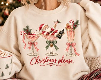 Könnte beinhalten: Ein cremefarbenes Sweatshirt mit Rundhalsausschnitt und einer Grafik von drei Cocktailgläsern, die mit weihnachtlichen Elementen wie einem liegenden Weihnachtsmann, einem Lebkuchenmann und Kirschen verziert sind. Der Text "Christmas please" steht in Rot darunter.