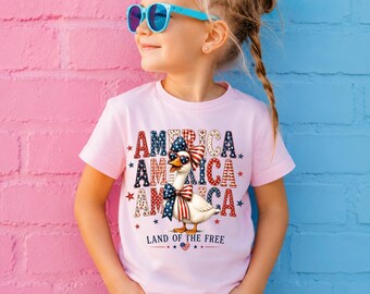 Könnte beinhalten: Ein hellrosa T-Shirt mit kurzen Ärmeln und einer Grafik einer weißen Gans, die eine rot-weiß-blaue Schleife und herzförmige Sonnenbrillen trägt. Der Text lautet AMERICA in einem patriotischen Muster sowie LAND OF THE FREE mit einer Flagge.