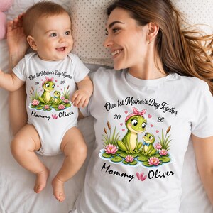 Könnte beinhalten: Weißes T-Shirt und Baby-Body mit passendem Frosch-Motiv. Das Design enthält den Text "Our 1st Mother's Day Together 20 26" und "Mommy Oliver". Die Frosch-Illustration zeigt eine rosa Schleife und Seerosenblätter.