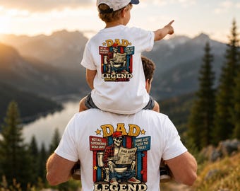 Könnte beinhalten: Weiße T-Shirts mit einem Grafikdesign mit dem Wort "DAD", einem Skelett, das eine Zeitung auf einer Toilette liest, und dem Text "LEGEND SINCE DAY 1". Ein Kind sitzt auf den Schultern eines Mannes und zeigt auf die Berge.