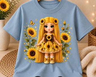 Könnte beinhalten: Ein hellblaues T-Shirt mit kurzen Ärmeln und einem zentralen Motiv einer Puppe mit langem gelben Haar, die ein gelbes Kleid mit Sonnenblumenmuster trägt. Die Puppe ist von Sonnenblumen und einem kleinen Schmetterling umgeben.