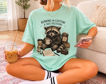 Könnte beinhalten: Hellgrünes T-Shirt mit dem Text "RUNNING ON CAFFEINE & BAD DECISIONS" und einer Waschbär-Grafik. Der Waschbär hält eine Kaffeetasse. Das Shirt wird mit weißen Socken mit schwarzen Streifen getragen.