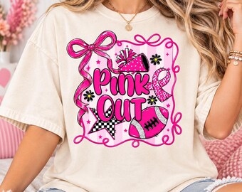 Könnte beinhalten: Ein cremefarbenes T-Shirt mit einer Grafik, die den Text Pink Out in fetten rosa Buchstaben zeigt. Das Design umfasst eine große rosa Schleife, ein Megafon, einen Football, einen karierten Stern und eine Schleife zur Krebsvorsorge.