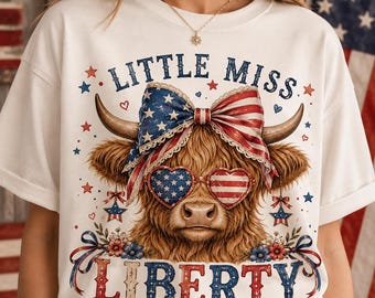 Könnte beinhalten: Ein weißes T-Shirt mit einer Grafik eines braunen Hochlandrindes, das eine herzförmige Brille mit US-Flaggenmuster und eine große patriotische Schleife trägt. Der Text lautet LITTLE MISS LIBERTY, umgeben von kleinen roten und blauen Sternen.