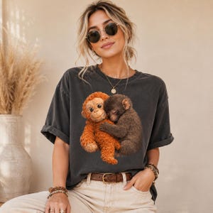 Matching oversized graphic t-shirts featuring a cute baby monkey and plush toy design. Lifestyle mockup with man and woman wearing vintage Comfort Colors apparel in neutral tones.
