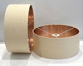 May include: Two beige fabric lampshades with copper-colored interiors. The lampshades are cylindrical, with a textured fabric exterior. One shade is upright, while the other lies on its side, showcasing the copper lining.
