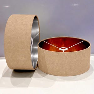 May include: Two cylindrical lamp shades with a textured beige fabric exterior. One shade stands upright, revealing a metallic copper interior, while the other rests on its side, showing a silver-toned interior. Both feature white wire frames.
