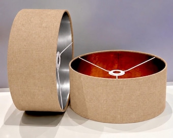 May include: Two cylindrical lamp shades with a textured beige fabric exterior. One shade stands upright, revealing a metallic copper interior, while the other rests on its side, showing a silver-toned interior. Both feature white wire frames.