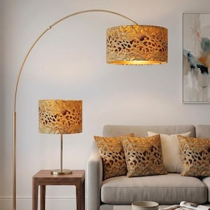 Può includere: Una lampada da terra ad arco dorata e una lampada da tavolo abbinata con paralumi a fantasia. I paralumi e i cuscini decorativi presentano un design oro e marrone. Sono visibili anche un divano grigio chiaro e un tavolino quadrato in legno.