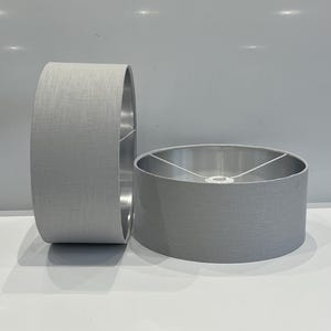 May include: Two grey fabric lampshades. One is upright, the other is on its side. The shades have a cylindrical shape with a textured fabric exterior and a silver interior. The lampshades are on a white surface.