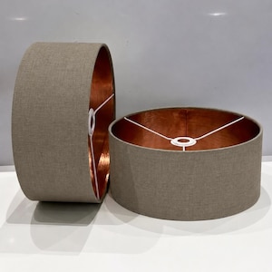May include: Two cylindrical lamp shades featuring a textured light brown fabric exterior and a metallic copper interior lining. Each shade includes a white wire frame structure with a central ring fitting for attachment to a lamp base fixture.