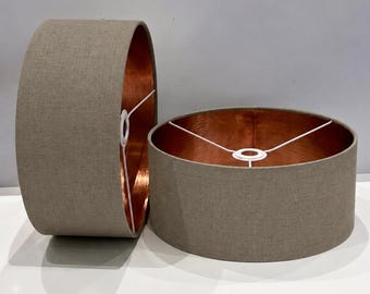 May include: Two cylindrical lamp shades featuring a textured light brown fabric exterior and a metallic copper interior lining. Each shade includes a white wire frame structure with a central ring fitting for attachment to a lamp base fixture.